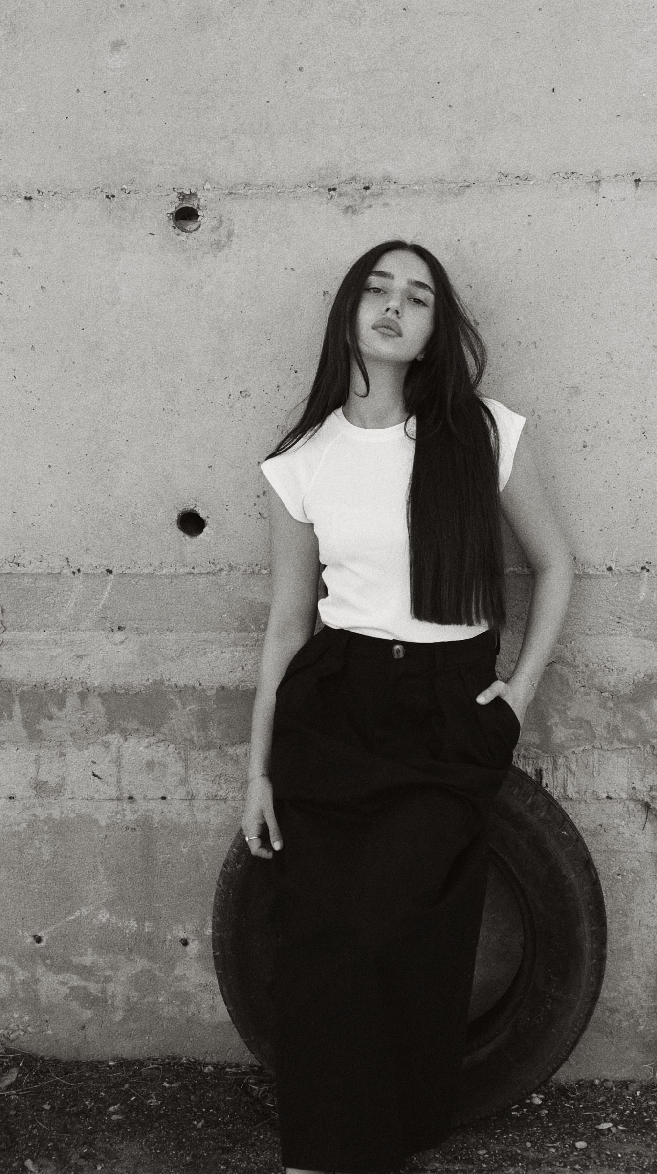 Black-and-white HILI RAZ campaign portrait: white cap-sleeve tee and high-waisted black tailoring against a concrete wall