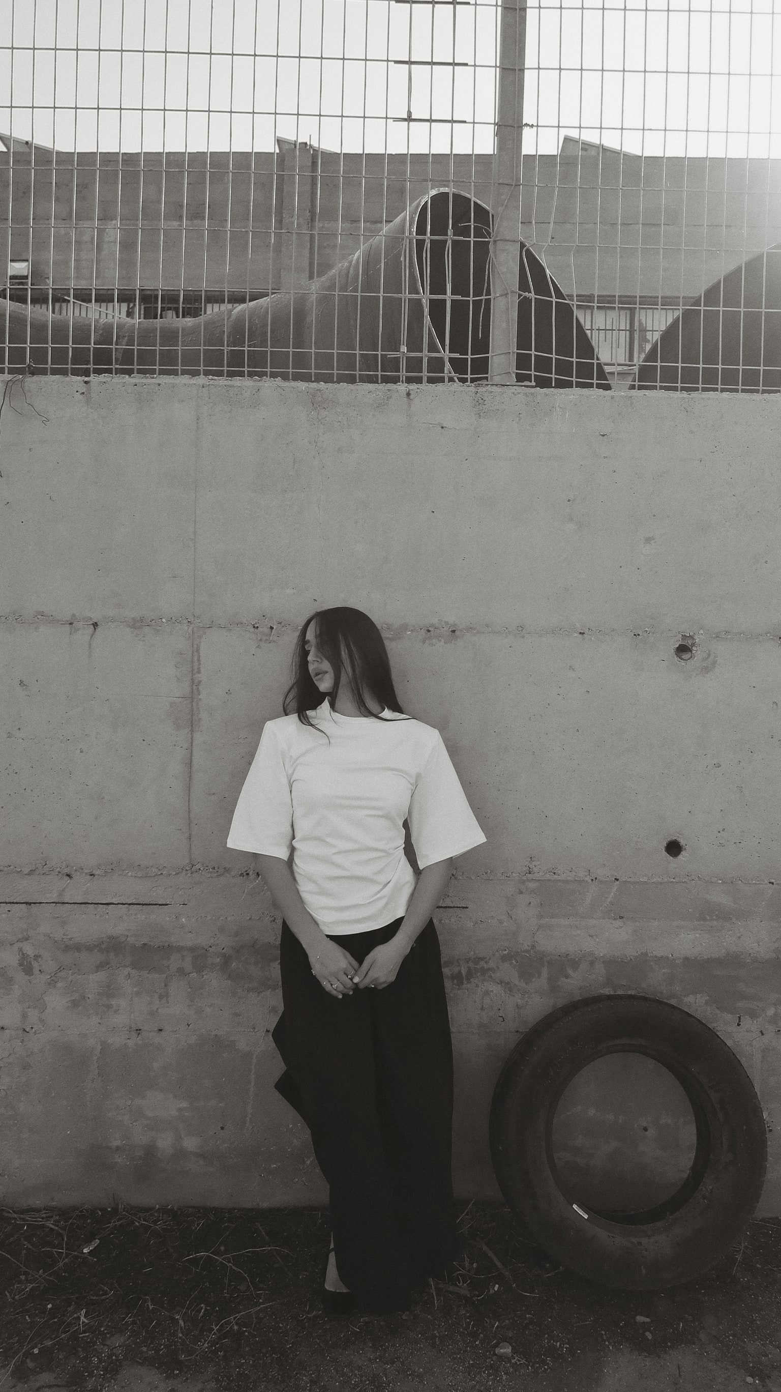 Black-and-white HILI RAZ campaign photo: white boxy tee and wide black tailoring beside a tyre at a concrete wall